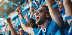 Mostbet | Nadšení fotbaloví fanoušci v modrých dresech oslavují gól na stadionu